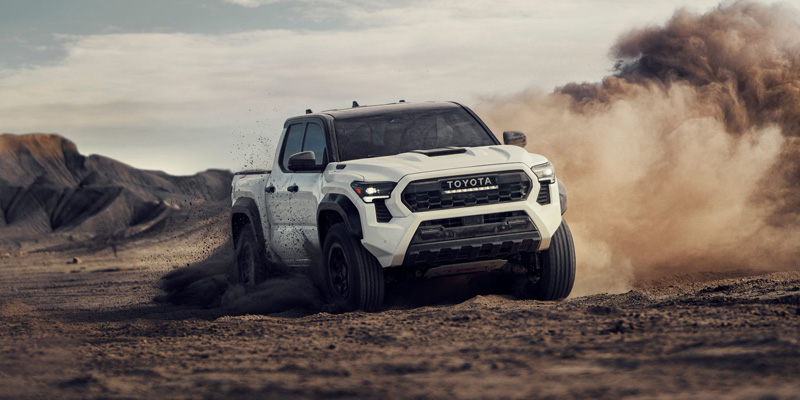 White Toyota Tacoma pickup truck driving off-road through a dusty desert trail.